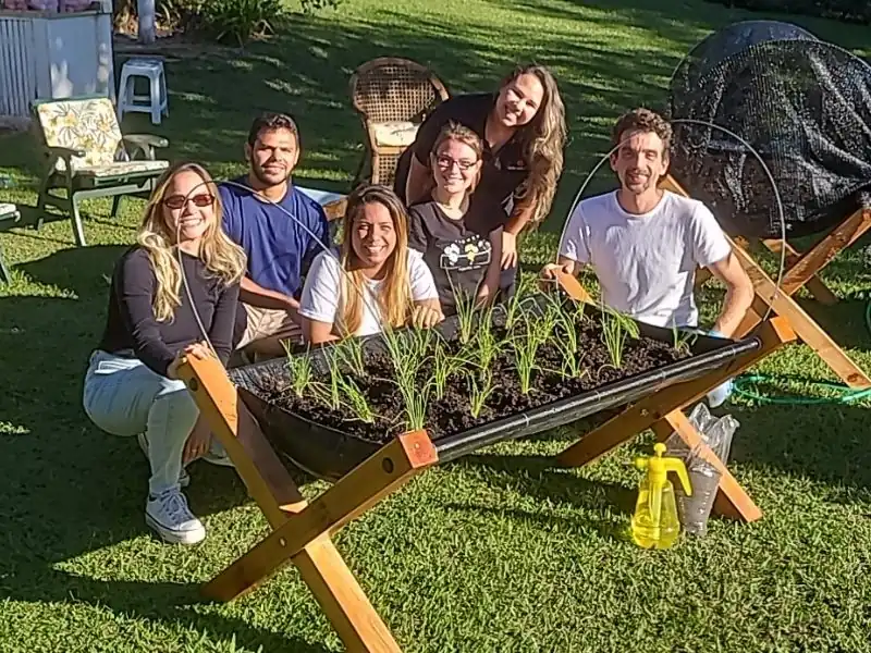 Equipe da Rede Educare e Jardim de Comer sorrindo atrás de um canteiro elevado repleto de cebolinhas verdes e saudáveis.