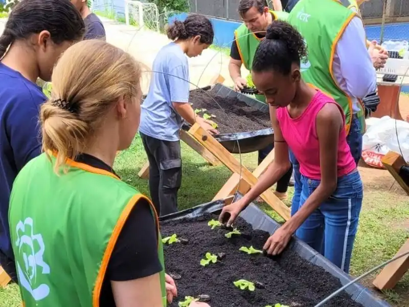 Vista de alunos plantando junto com voluntária mudas de hortaliças em canteiro de horta elevado em escola.