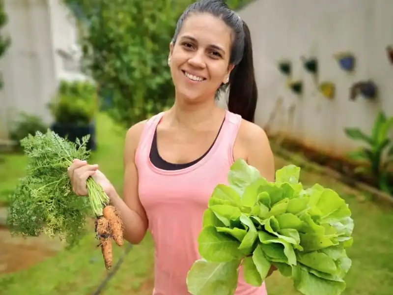 Moradora sorridente segurando cenouras e alface fresca colhidas na horta comunitária.
