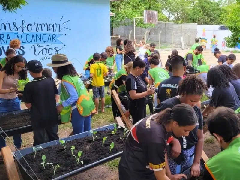 Foto panorâmica do pátio da escola com diversos alunos e voluntários trabalhando na manutenção e plantio de hortas elevadas.
