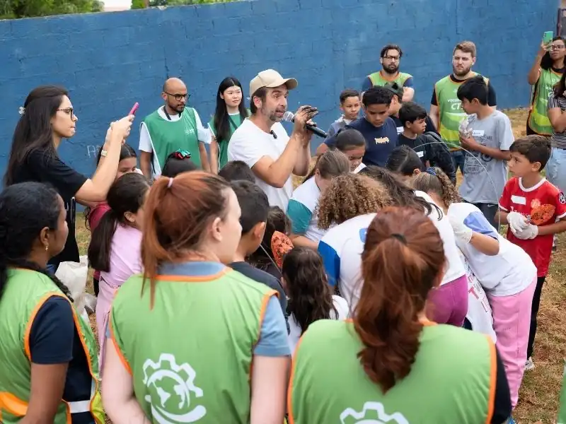 Rodolfo Rodrigues fala ao microfone para um grupo de crianças e voluntários em volta de uma horta, durante atividade de educação ambiental.
