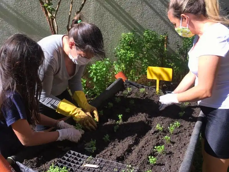 Duas mulheres e uma criança plantando mudas juntas em canteiro de horta escolar/condomínio.