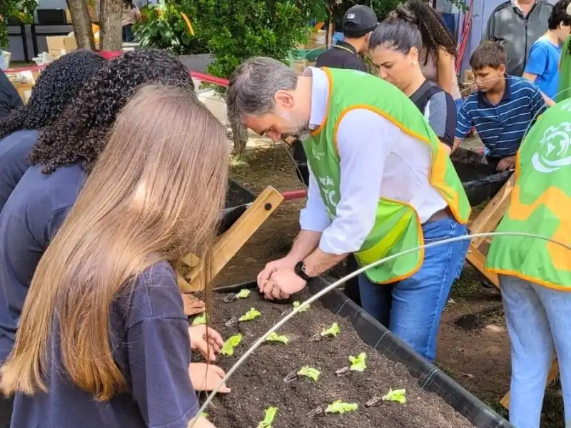 Voluntário usando colete verde do projeto, ensina alunos a plantarem mudas em uma horta elevada em ambiente escolar.