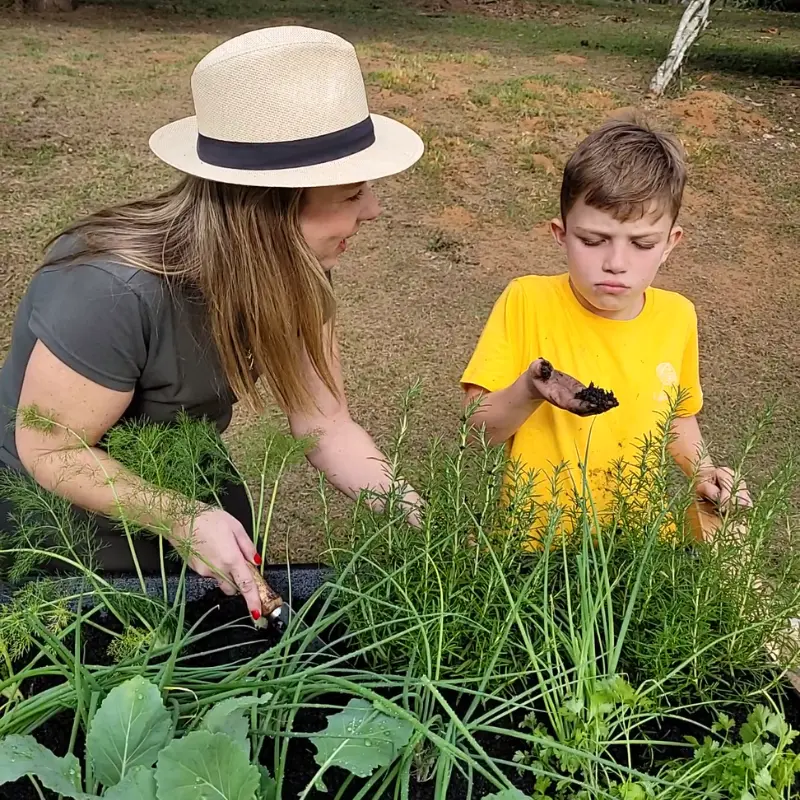 Mãe e filho cuidando da horta suspensa do Jardim de Comer, criança segurando terra adubada no canteiro elevado.