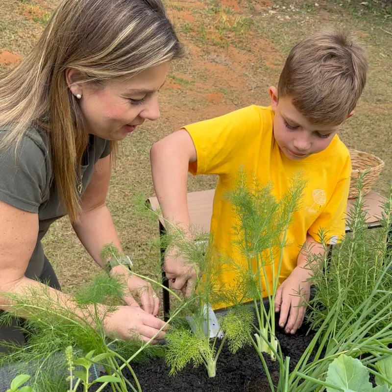 Mãe e filho plantando ervas na horta suspensa do Jardim de Comer, preparando a terra no canteiro elevado ao ar livre.