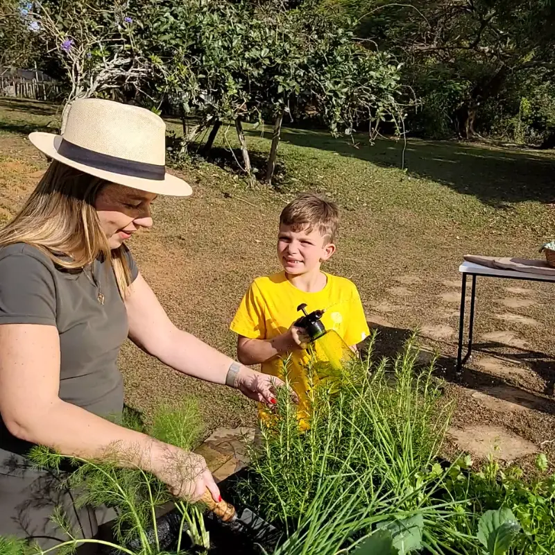 Mãe com chapéu cuidando da horta suspensa do Jardim de Comer enquanto o filho segura borrifador, cultivo de temperos em casa.