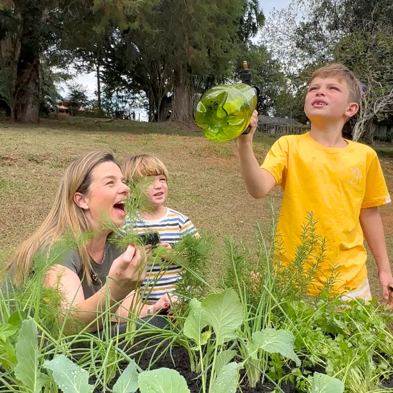 Família colhendo e regando a horta suspensa do Jardim de Comer; criança com borrifador e canteiro elevado com verduras e temperos.