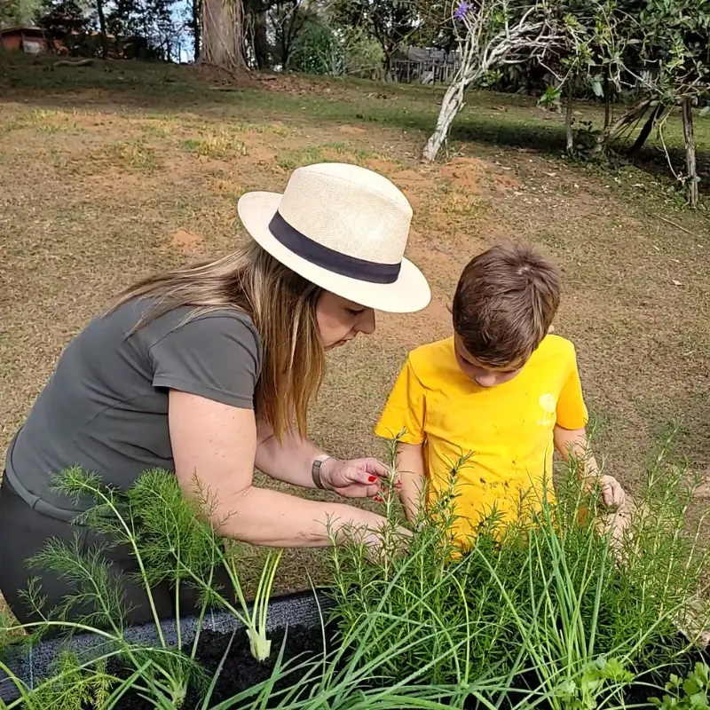 Mãe e filho fazendo manutenção na horta suspensa do Jardim de Comer, ajustando mudas e ervas no canteiro elevado no jardim.
