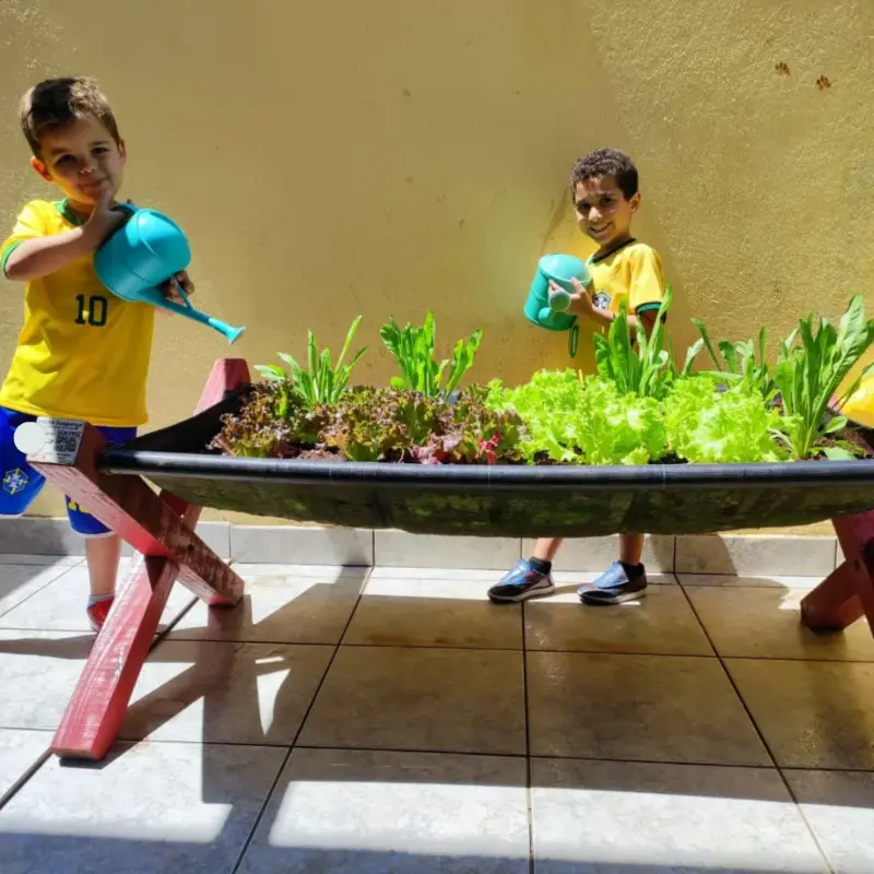 Dois alunos regando canteiro elevado na horta escolar, cultivo de alface e hortaliças em ambiente escolar.