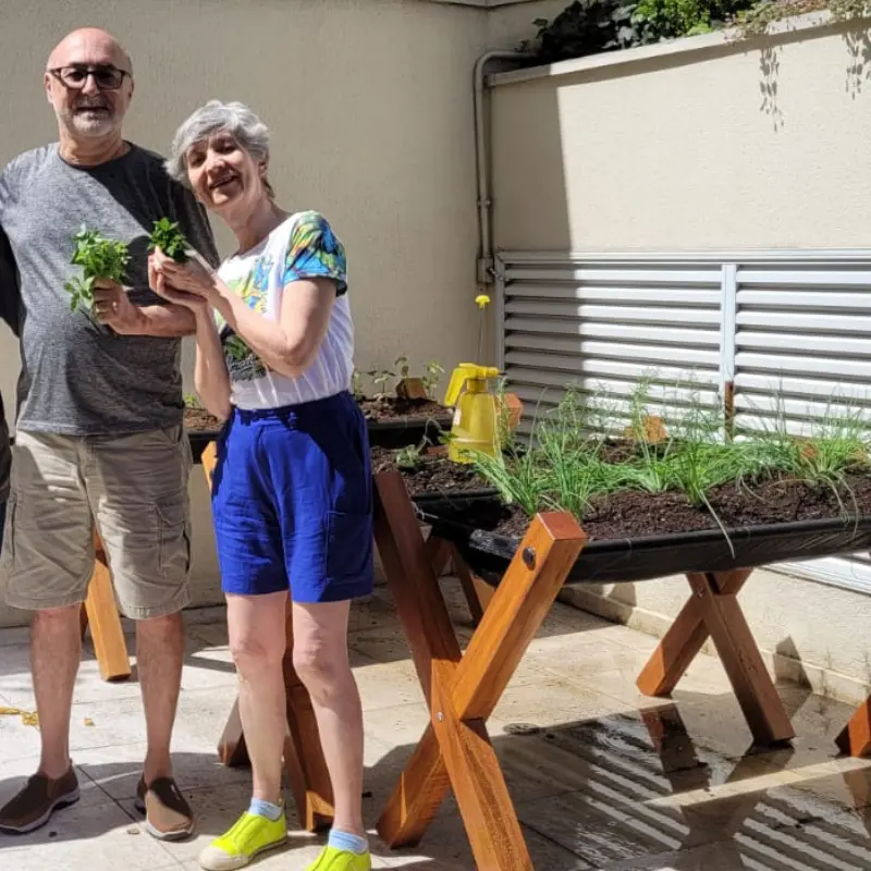 Casal mostrando a colheita em um cantinho de ervas e temperos, em espaço residencial ensolarado.