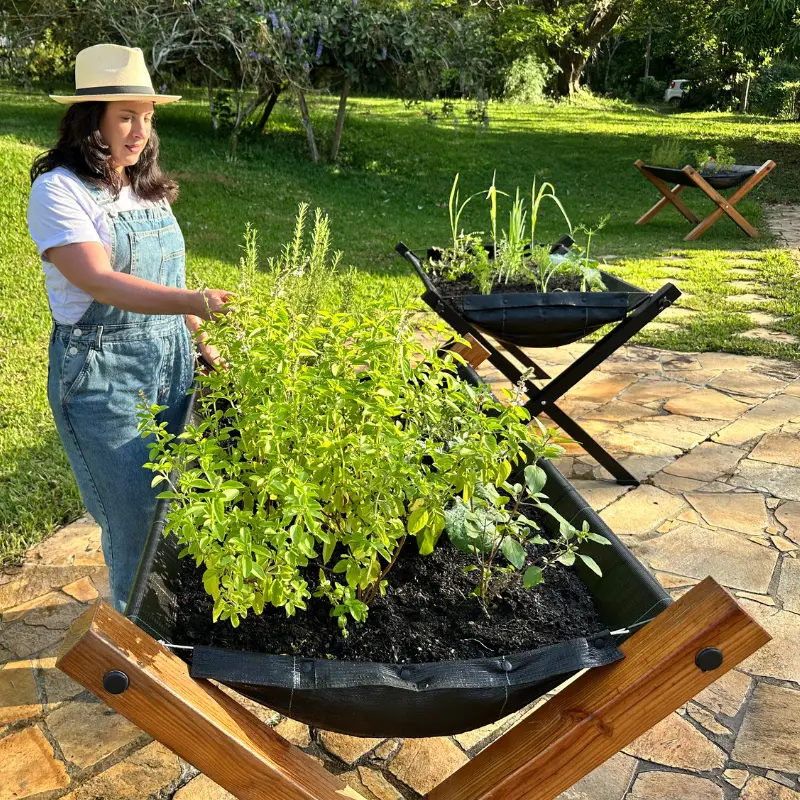 Mulher observando e colhendo em um cantinho de ervas e temperos, em área externa com mais de uma estrutura.